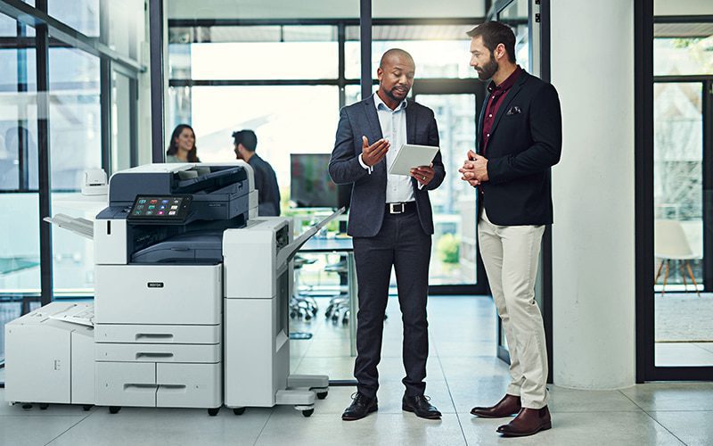 Twee mannen in een kantooromgeving die iets bespreken op een tablet naast een Xerox® AltaLink® C8200 Series Colour Multifunction Printer