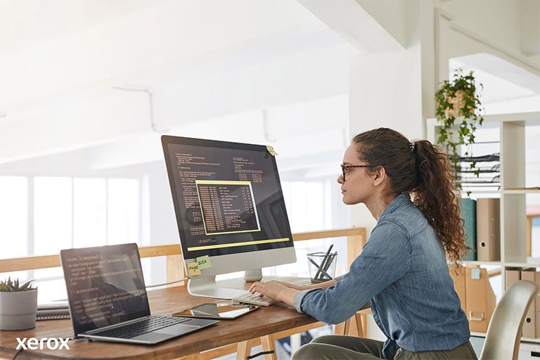 Een vrouw aan haar bureau, bezig met een computer en laptop, demonstreert productiviteit in een moderne werkruimte.