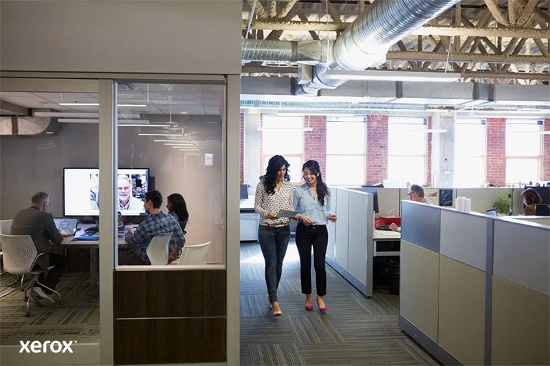 Twee vrouwen lopen door een moderne open kantoorruimte, in gesprek en omringd door werkstations.